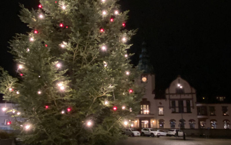 Julgran med tända ljus i förgrunden och Rådhuset i bakgrunden på Stortorget i Karlshamn. Det är mörkt och kallt och vinter.