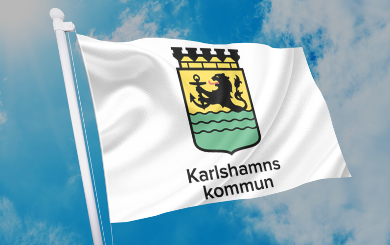 Karlshamns kommunflagga med Karlshamns vapen på vit botten fladdrar i vinden mot en blå himmel.